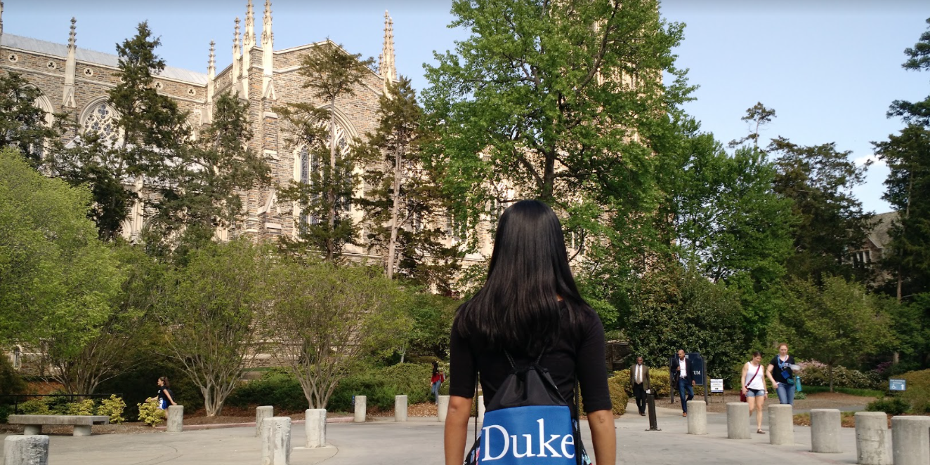 A girl with her back to the camera.
      She is wearing a dark blue bag that says 'Duke' on it and is looking at 
      a tall building in the distance.